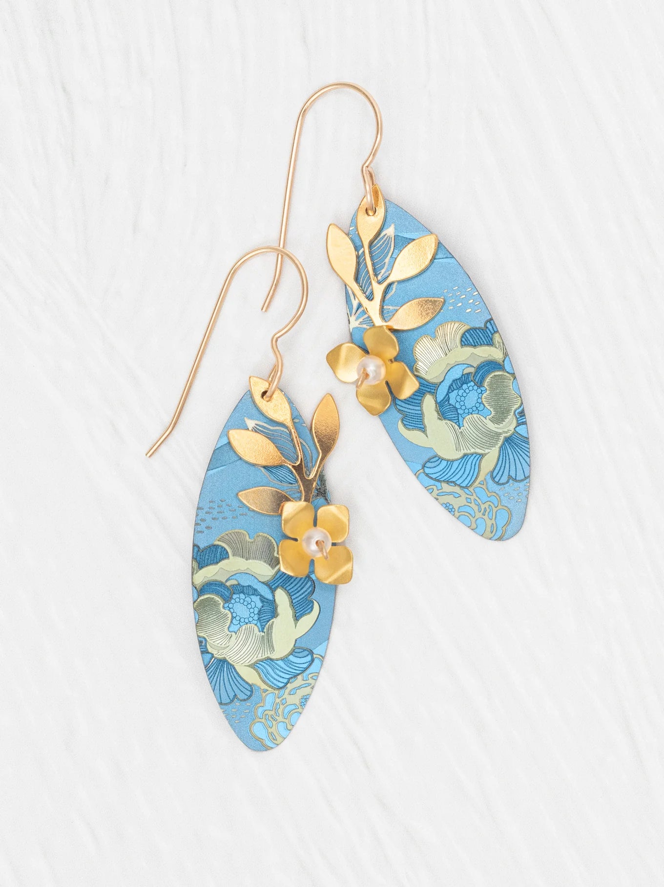 The Alyssa Sky Blue Earrings by Holly Yashi feature elongated blue ovals with gold floral patterns, leaf and flower accents, and hand-colored niobium, shown on a white textured background.