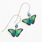 The Green Flash Petite Bella Butterfly Earrings from Holly Yashi feature vibrant blue and green butterfly wings with brown edges. They elegantly hang from sterling silver hooks adorned with small blue beads, showcased beautifully against a crisp white background.