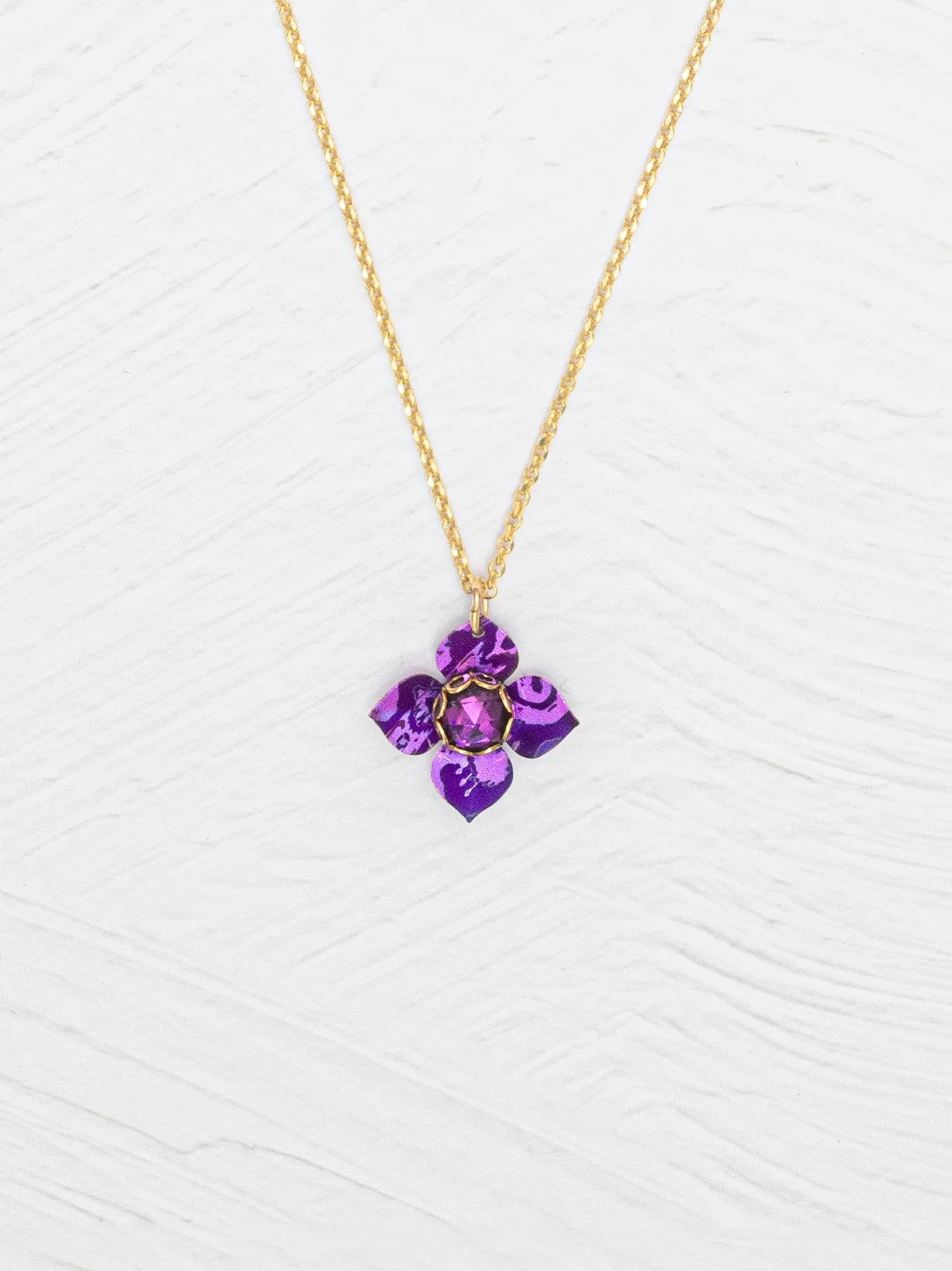 The Petite Fleur Royal Purple Necklace by Holly Yashi features a gold chain and a purple flower-shaped Austrian crystal pendant, shown on a white textured background.