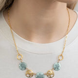 A model in a white top features the Sage Oceana Necklace from Holly Yashi Jewelry, with a gold chain adorned with round turquoise disks and small gold-plated shells, elegantly enhancing the neckline.