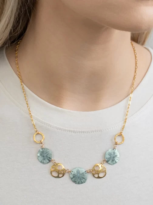 A model in a white top features the Sage Oceana Necklace from Holly Yashi Jewelry, with a gold chain adorned with round turquoise disks and small gold-plated shells, elegantly enhancing the neckline.