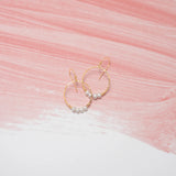 The Rosa Gold and Pearls Hoop Earrings by Holly Yashi are elegantly displayed on a surface with pink and white brush strokes, adding a sophisticated touch.