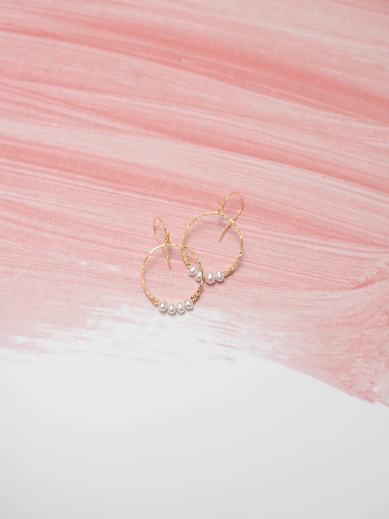 The Rosa Gold and Pearls Hoop Earrings by Holly Yashi are elegantly displayed on a surface with pink and white brush strokes, adding a sophisticated touch.