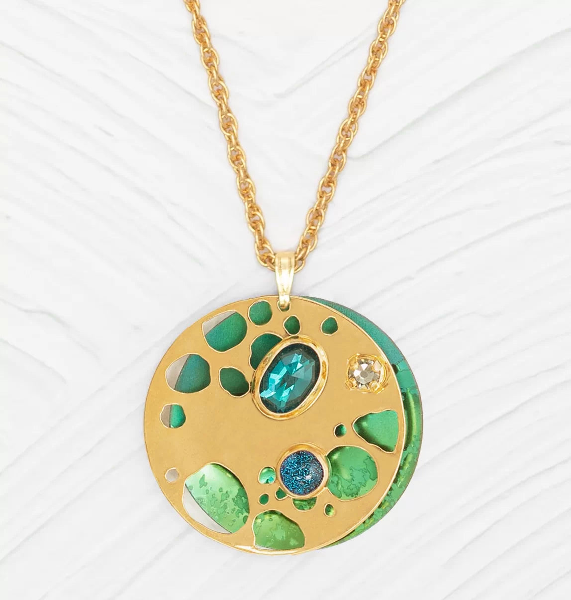 The Ocean Depths Teal Necklace by Holly Yashi showcases a gold pendant with a round disc embellished with blue and green gems, mirroring ocean colors. Its twisted gold chain adds elegance against a textured white surface, enhanced by intricate cut-out designs.