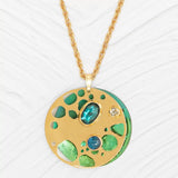 The Ocean Depths Teal Necklace by Holly Yashi showcases a gold pendant with a round disc embellished with blue and green gems, mirroring ocean colors. Its twisted gold chain adds elegance against a textured white surface, enhanced by intricate cut-out designs.