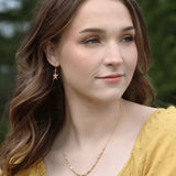 A woman with long brown hair wears a yellow top and Carmel Seashore Peach Starfish Earrings by Holly Yashi. Shes also adorned with a star pendant necklace. With green trees behind her, she gazes to the side, smiling slightly.