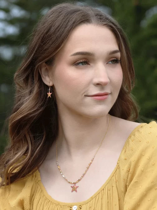 A woman with long brown hair wears a yellow top and Carmel Seashore Peach Starfish Earrings by Holly Yashi. Shes also adorned with a star pendant necklace. With green trees behind her, she gazes to the side, smiling slightly.