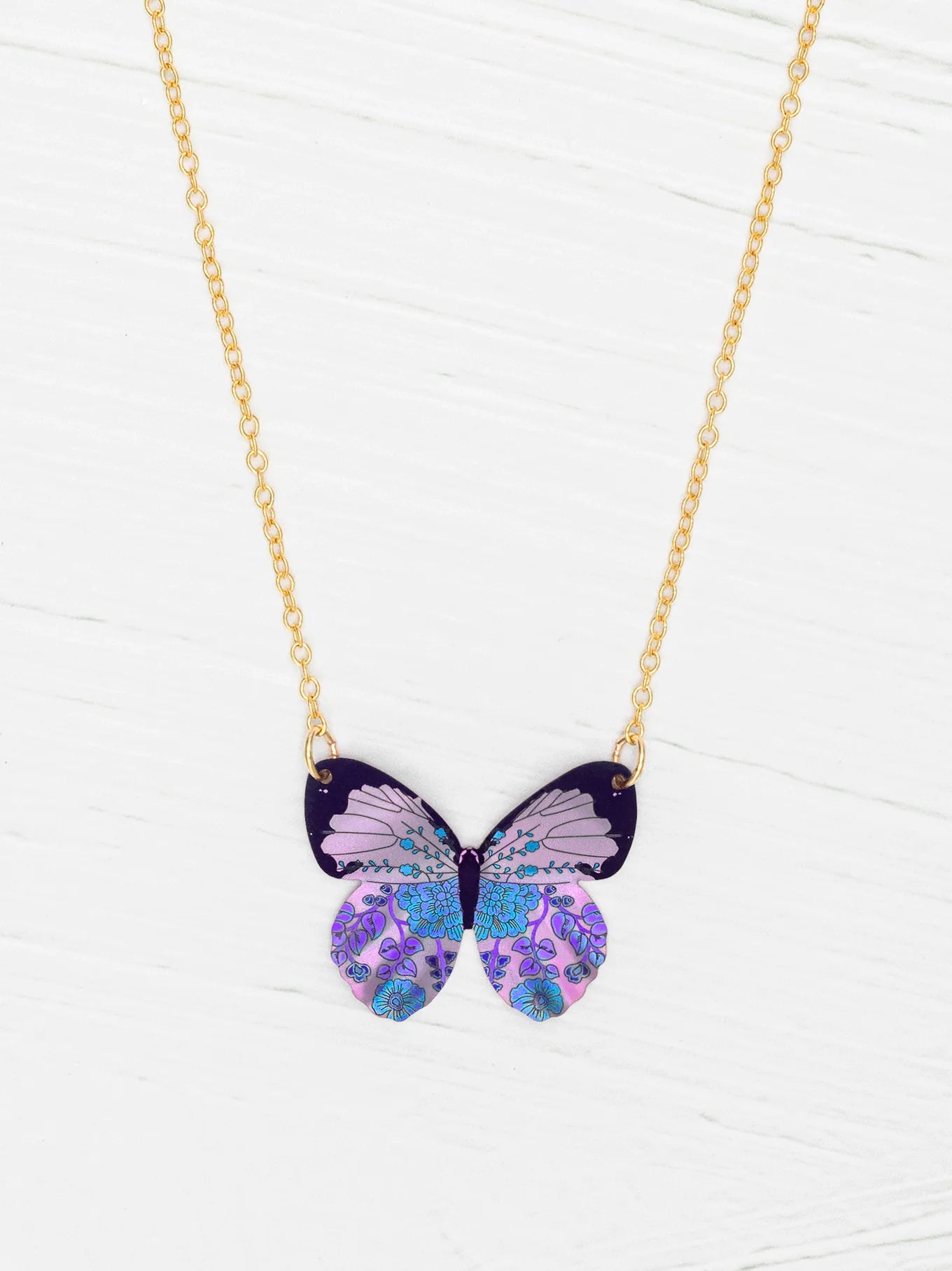 The Blooming Butterfly Radiant Purple Necklace by Holly Yashi features an iridescent niobium butterfly pendant with purple, blue, and lavender patterns on a gold adjustable chain, shown on a white wooden surface.
