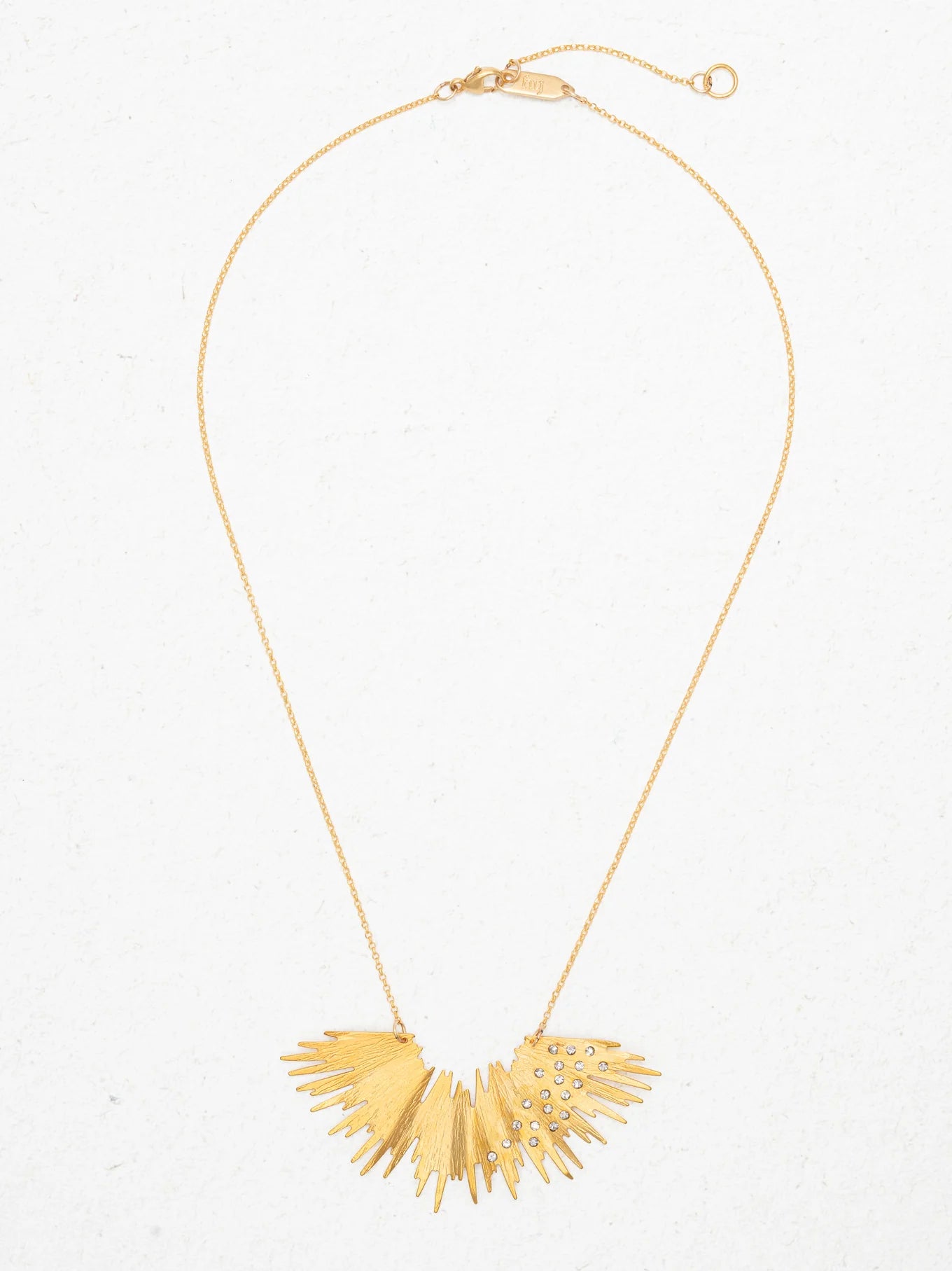 The Holly Yashi Aster Necklace-Gold Necklace features a delicate chain and a sunburst-shaped pendant with textured rays and sparkling crystal stones, beautifully displayed on a light background.