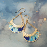 The Selena/Midnight Blue earrings by Holly Yashi feature gold crescent moons, blue and white abstract designs, rhinestones, and gold chain links. Hypoallergenic, they are shown on a blue watercolor-patterned background.