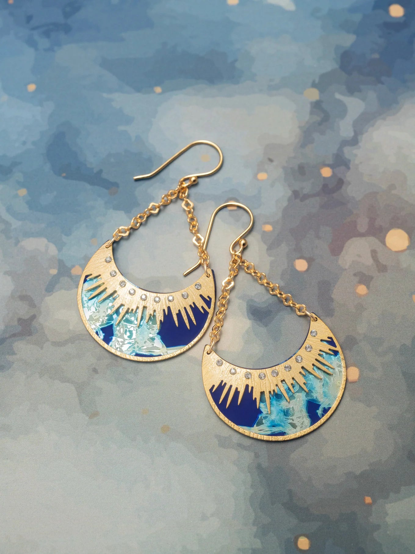 The Selena/Midnight Blue earrings by Holly Yashi feature gold crescent moons, blue and white abstract designs, rhinestones, and gold chain links. Hypoallergenic, they are shown on a blue watercolor-patterned background.