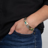 A person in blue jeans and a black shirt has their hand in their pocket, showcasing the Holly Yashi Gilded Petra Emerald Bracelet adorned with emerald green crystals and shimmering blue gemstones.