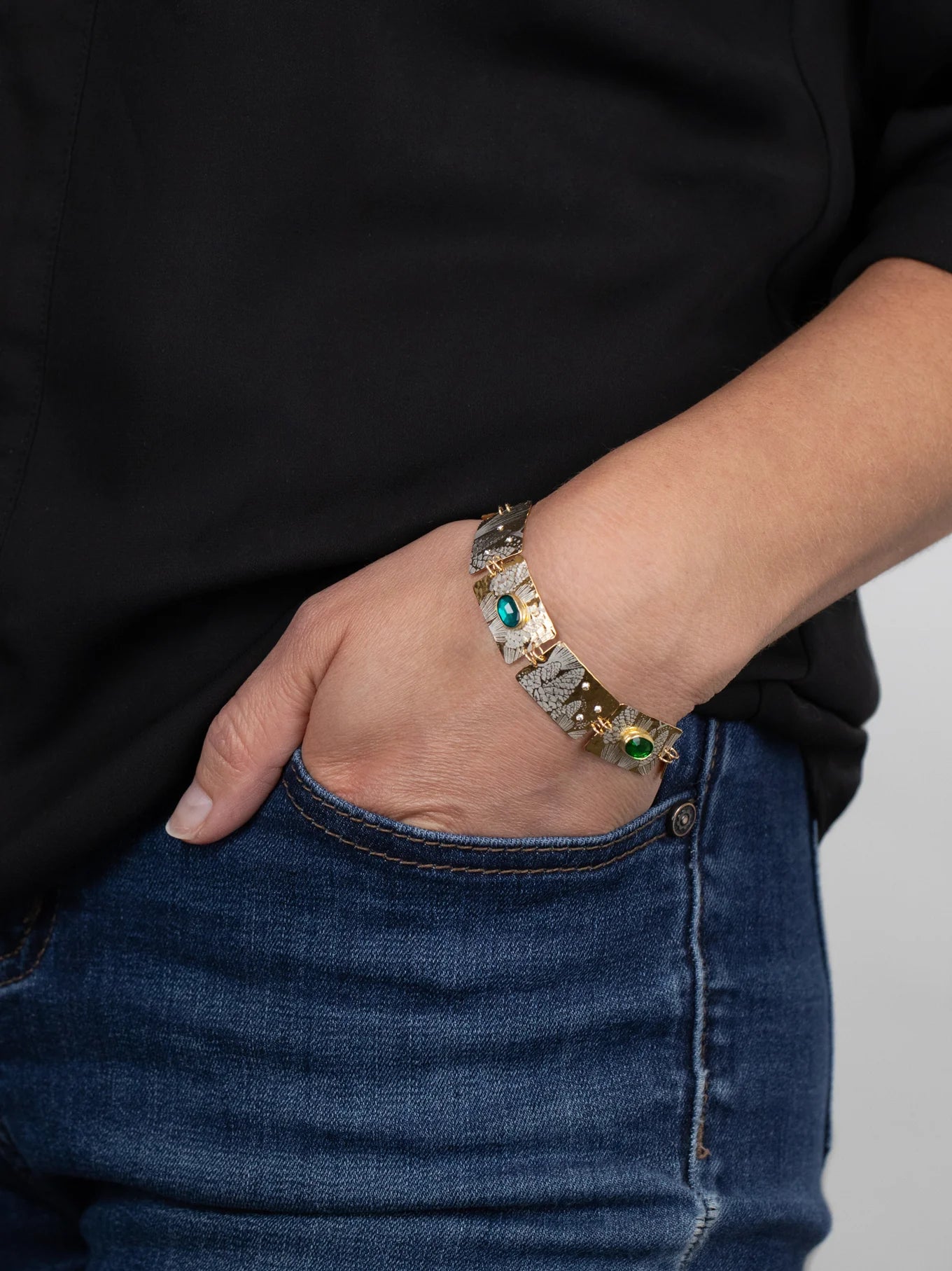 A person in blue jeans and a black shirt has their hand in their pocket, showcasing the Holly Yashi Gilded Petra Emerald Bracelet adorned with emerald green crystals and shimmering blue gemstones.