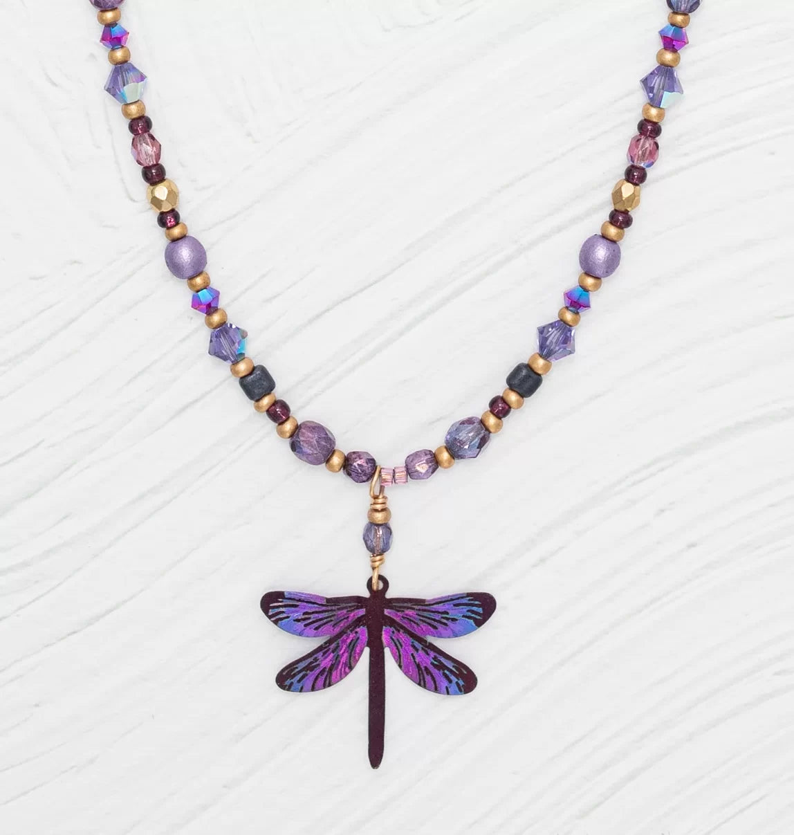 Introducing the Violet Skies Beaded Dragonfly Dreams Necklace from Holly Yashi Jewelry, adorned with 18k gold-plated purple, gold, and multicolored beads. It features a stunning central dragonfly pendant in shimmering purple and blue on a textured white background.