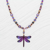 Introducing the Violet Skies Beaded Dragonfly Dreams Necklace from Holly Yashi Jewelry, adorned with 18k gold-plated purple, gold, and multicolored beads. It features a stunning central dragonfly pendant in shimmering purple and blue on a textured white background.