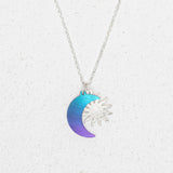 The Sun and Moon Calypso Necklace by Holly Yashi features a silver sun overlapping a crescent moon in a blue to purple gradient, displayed on a white textured background—ideal for celestial jewelry lovers.