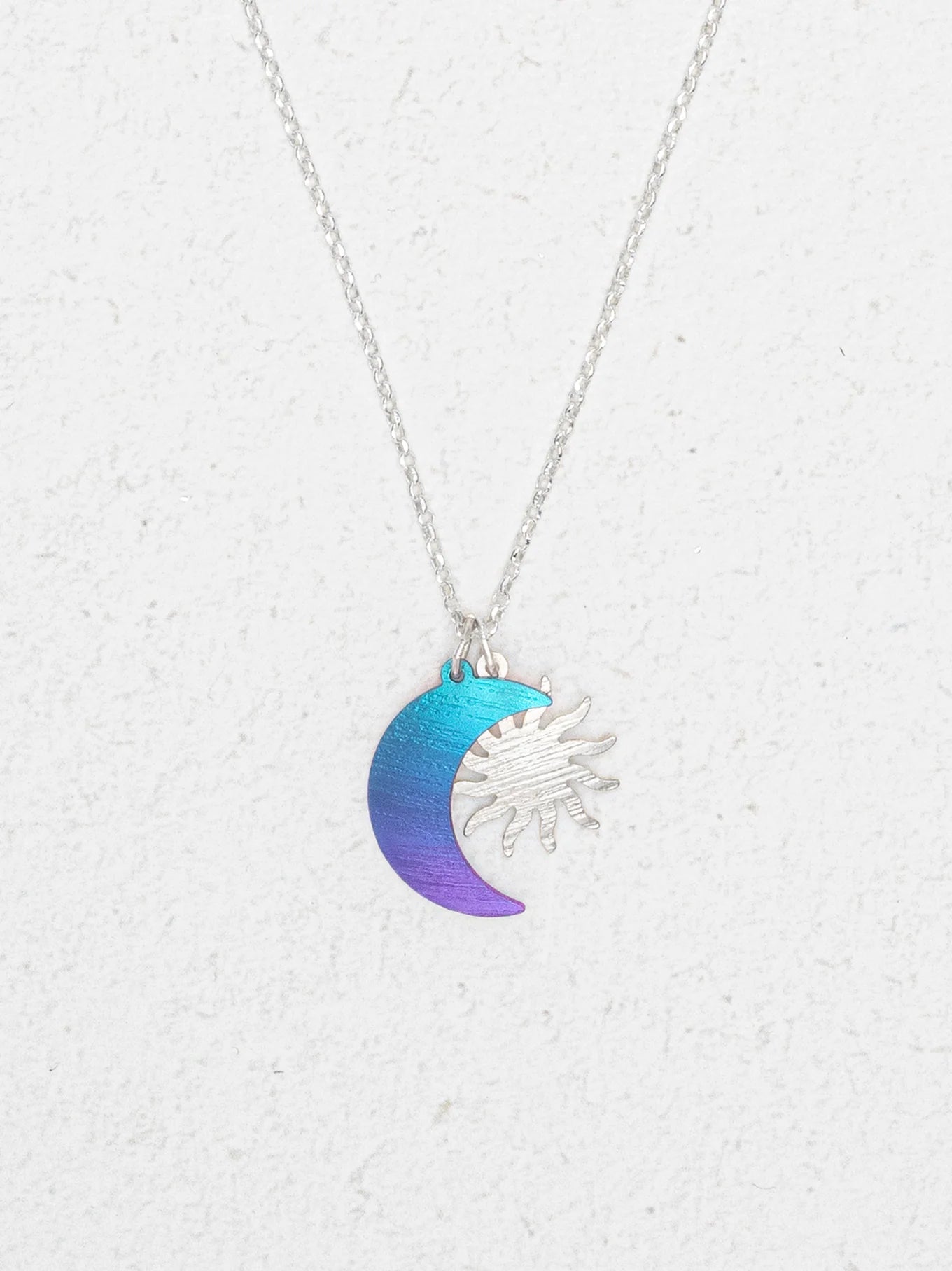 The Sun and Moon Calypso Necklace by Holly Yashi features a silver sun overlapping a crescent moon in a blue to purple gradient, displayed on a white textured background—ideal for celestial jewelry lovers.