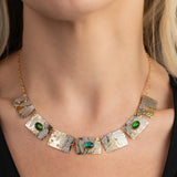 A close-up of a woman's neck wearing the Gilded Petra Emerald Necklace by Holly Yashi—a gold chain with textured rectangular plates and three emerald green crystals—paired with a black top for modern Victorian flair.