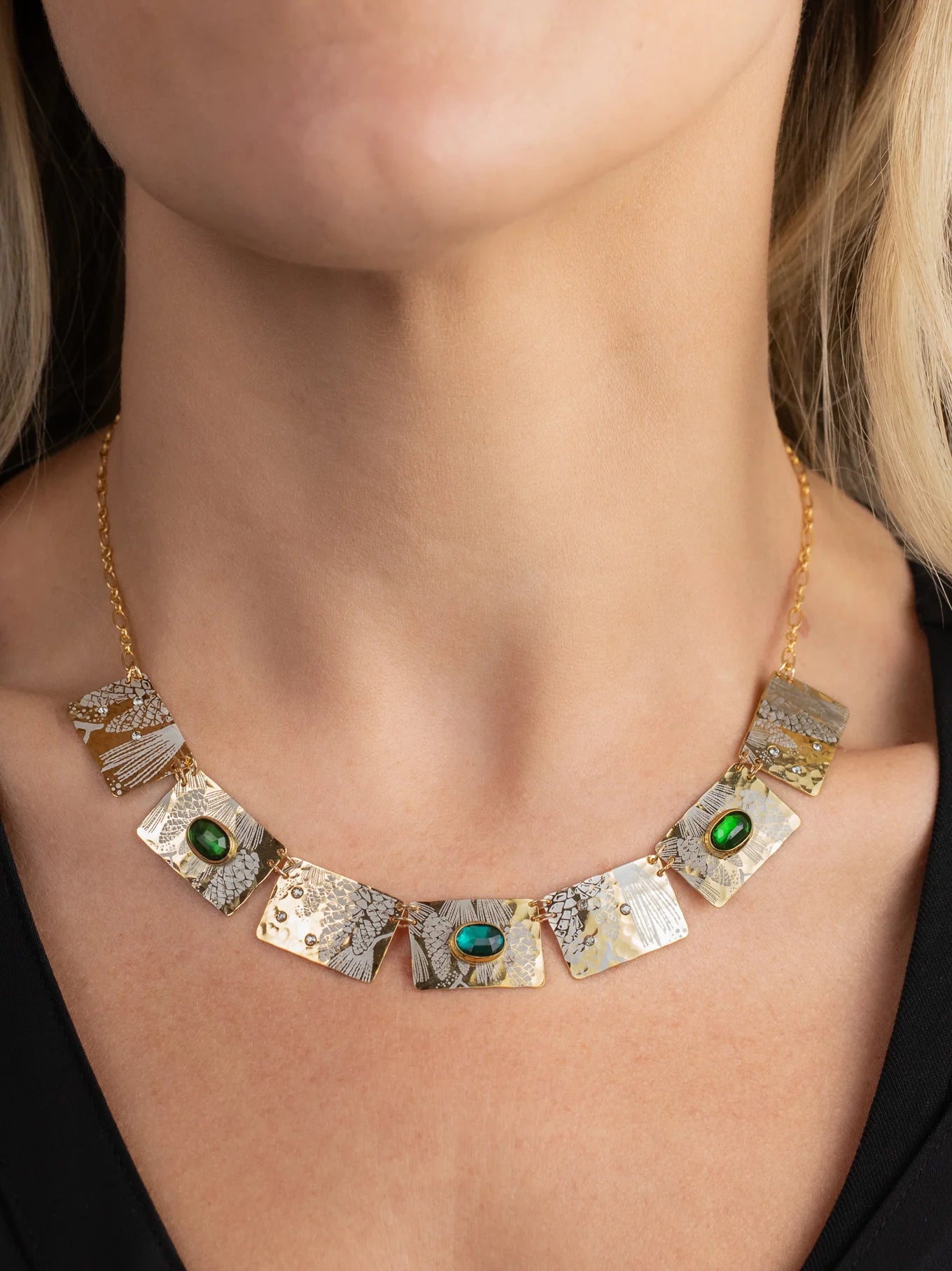 A close-up of a woman's neck wearing the Gilded Petra Emerald Necklace by Holly Yashi—a gold chain with textured rectangular plates and three emerald green crystals—paired with a black top for modern Victorian flair.