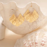 The Gold Ariel Earrings from Holly Yashi are elegantly showcased on white seashells against a neutral background, with their textured and reflective gold plating enhancing the overall elegance.