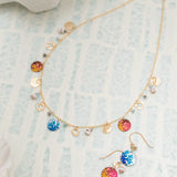 A delicate gold necklace and Holly Yashis Coral Reef Seashore Blue Earrings, featuring colorful round pendants with orange and blue sun motifs. The jewelry, crafted from hypoallergenic materials, is artfully displayed on a light patterned surface.
