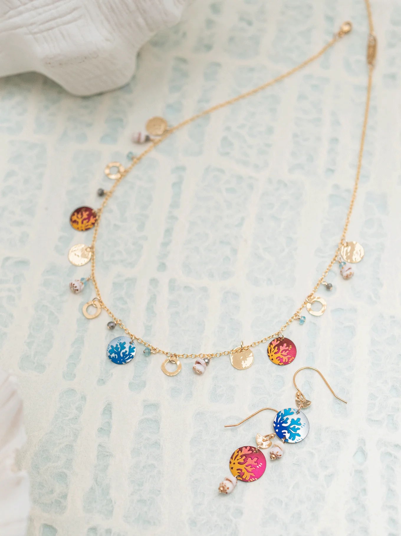 A delicate gold necklace and Holly Yashis Coral Reef Seashore Blue Earrings, featuring colorful round pendants with orange and blue sun motifs. The jewelry, crafted from hypoallergenic materials, is artfully displayed on a light patterned surface.