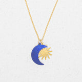 The Holly Yashi Sun and Moon Midnight Blue Necklace features a gold sun and a blue crescent moon pendant, displayed on a white textured background.