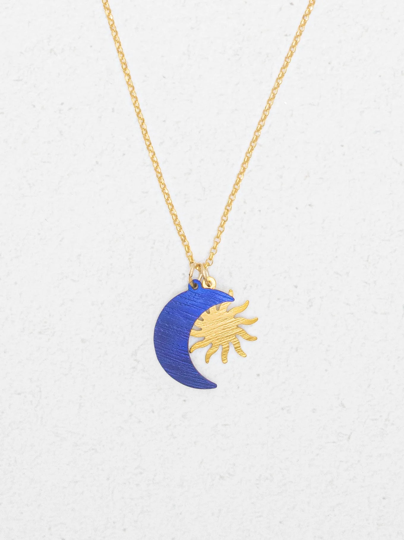 The Holly Yashi Sun and Moon Midnight Blue Necklace features a gold sun and a blue crescent moon pendant, displayed on a white textured background.