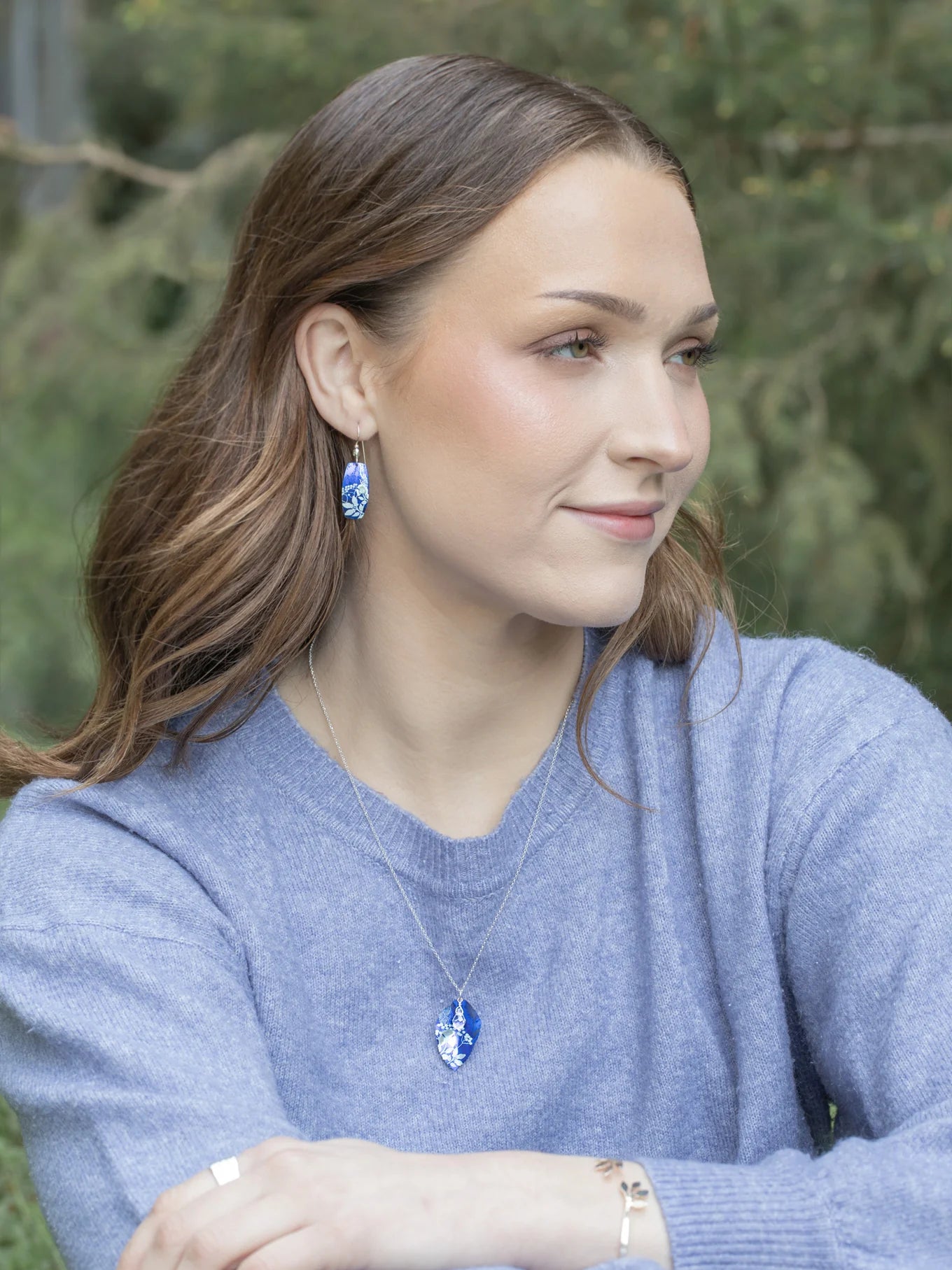 A woman with long brown hair, wearing a light blue sweater and blue and white drop earrings, sits outdoors with a gentle smile. She wears the Blooming Elm Indigo Necklace by Holly Yashi. Blurred green foliage is in the background.