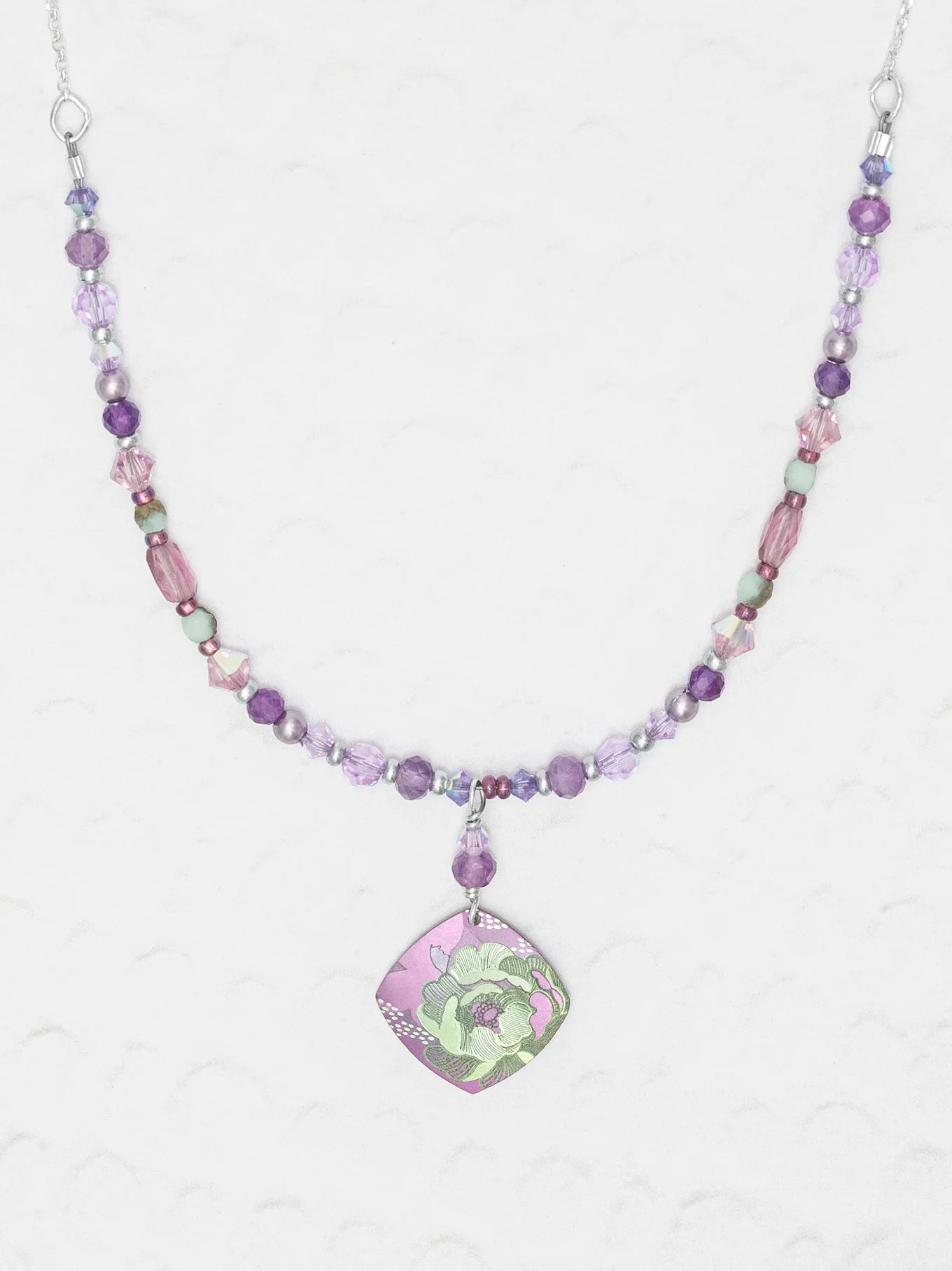 The Elena Beaded Necklace Violet by Holly Yashi features purple, pink, and green beads with a square floral green and purple pendant, shown on a textured white background.