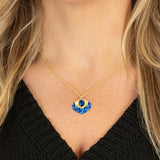 A woman wears the Holly Yashi Arabella Sapphire Blue Necklace, featuring an adjustable gold chain and a vibrant blue and gold glass stone pendant. She pairs it with a black V-neck top.