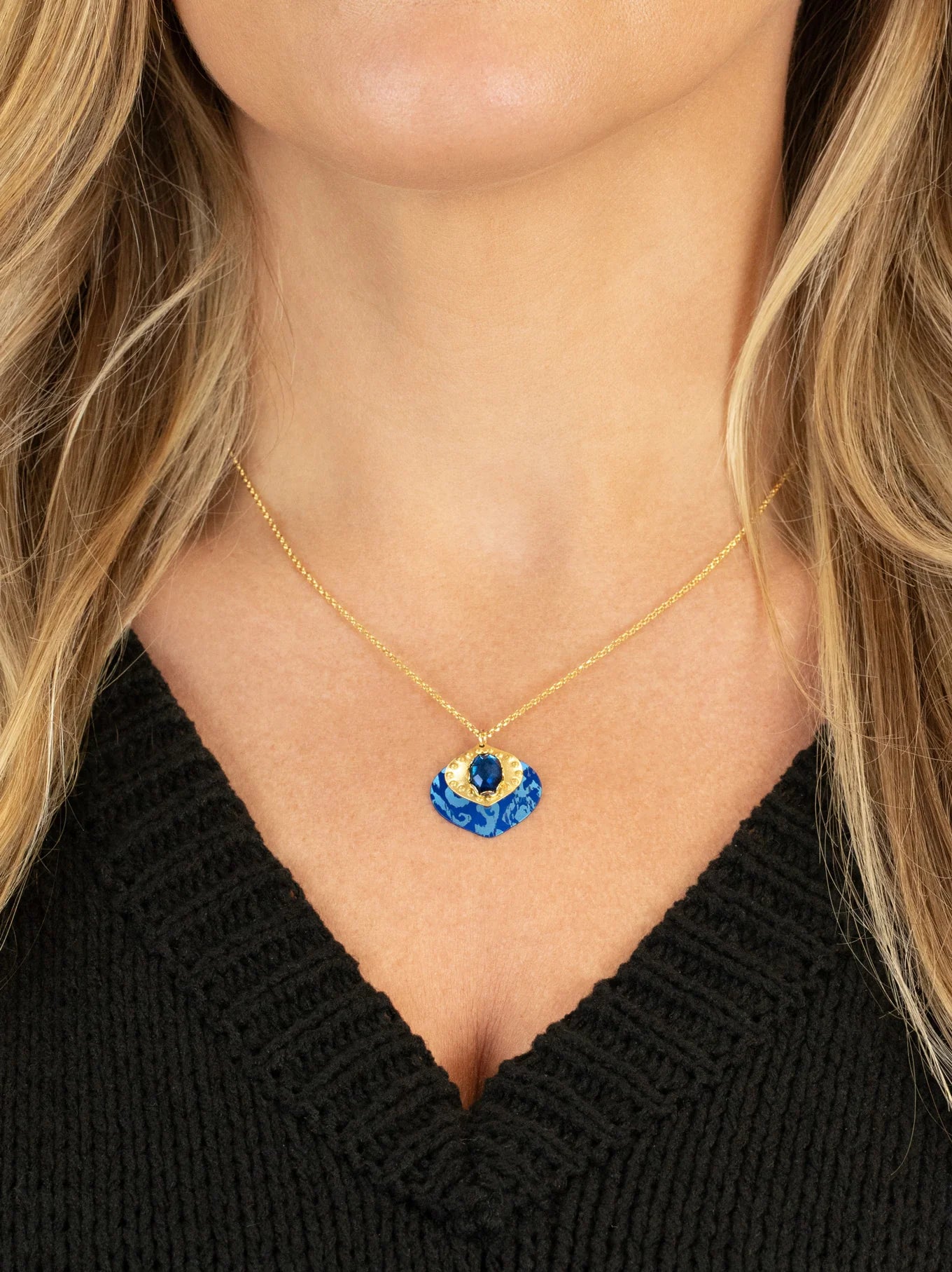 A woman wears the Holly Yashi Arabella Sapphire Blue Necklace, featuring an adjustable gold chain and a vibrant blue and gold glass stone pendant. She pairs it with a black V-neck top.