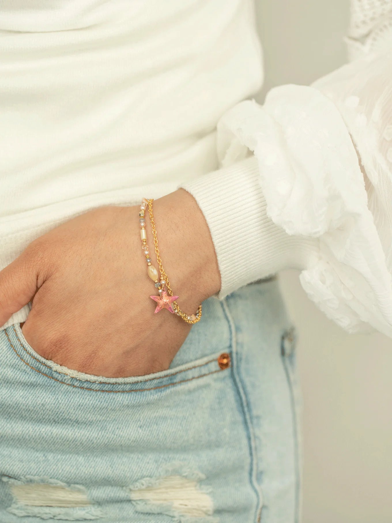 A person in light blue denim jeans, with a hand tucked in their pocket, showcases their wrist adorned with the Carmel Beaded Seashore Blue Starfish Bracelet from Holly Yashi Jewelry. The gold charm features a pink star, beautifully contrasting their white textured long-sleeve top.