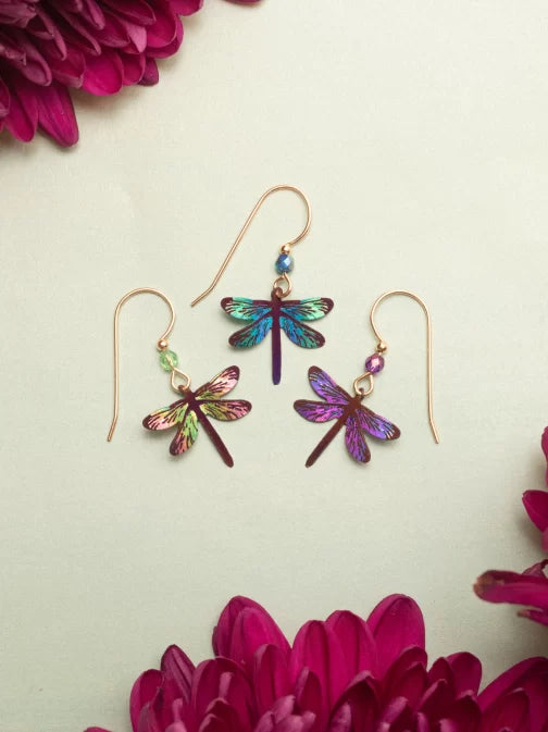 The Turquoise Dragonfly Dreams Earrings by Holly Yashi feature three iridescent dragonfly designs crafted from colorful niobium. Displayed against a light background with vibrant pink flowers, these hypo-allergenic earrings are perfect for sensitive skin and ideal for any jewelry enthusiast.