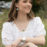 Amid the greenery, a brown-haired woman sits on grass in blue jeans and a white blouse, smiling softly. She gazes left while accessorized with Shelby Sage and Gold Earrings by Holly Yashi, a silver bracelet, layered necklaces, and Austrian crystal pearl accents.