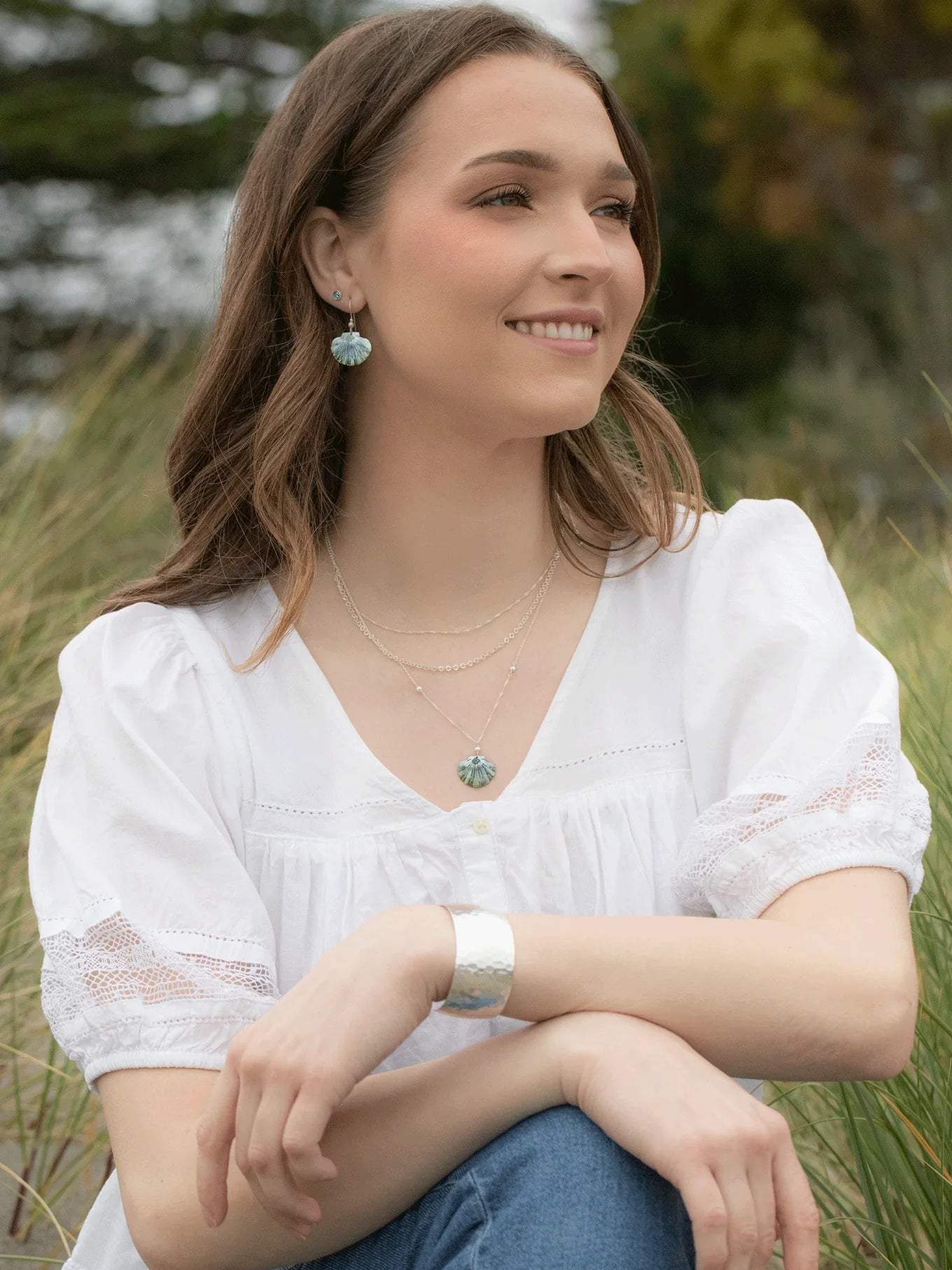 Amid the greenery, a brown-haired woman sits on grass in blue jeans and a white blouse, smiling softly. She gazes left while accessorized with Shelby Sage and Gold Earrings by Holly Yashi, a silver bracelet, layered necklaces, and Austrian crystal pearl accents.