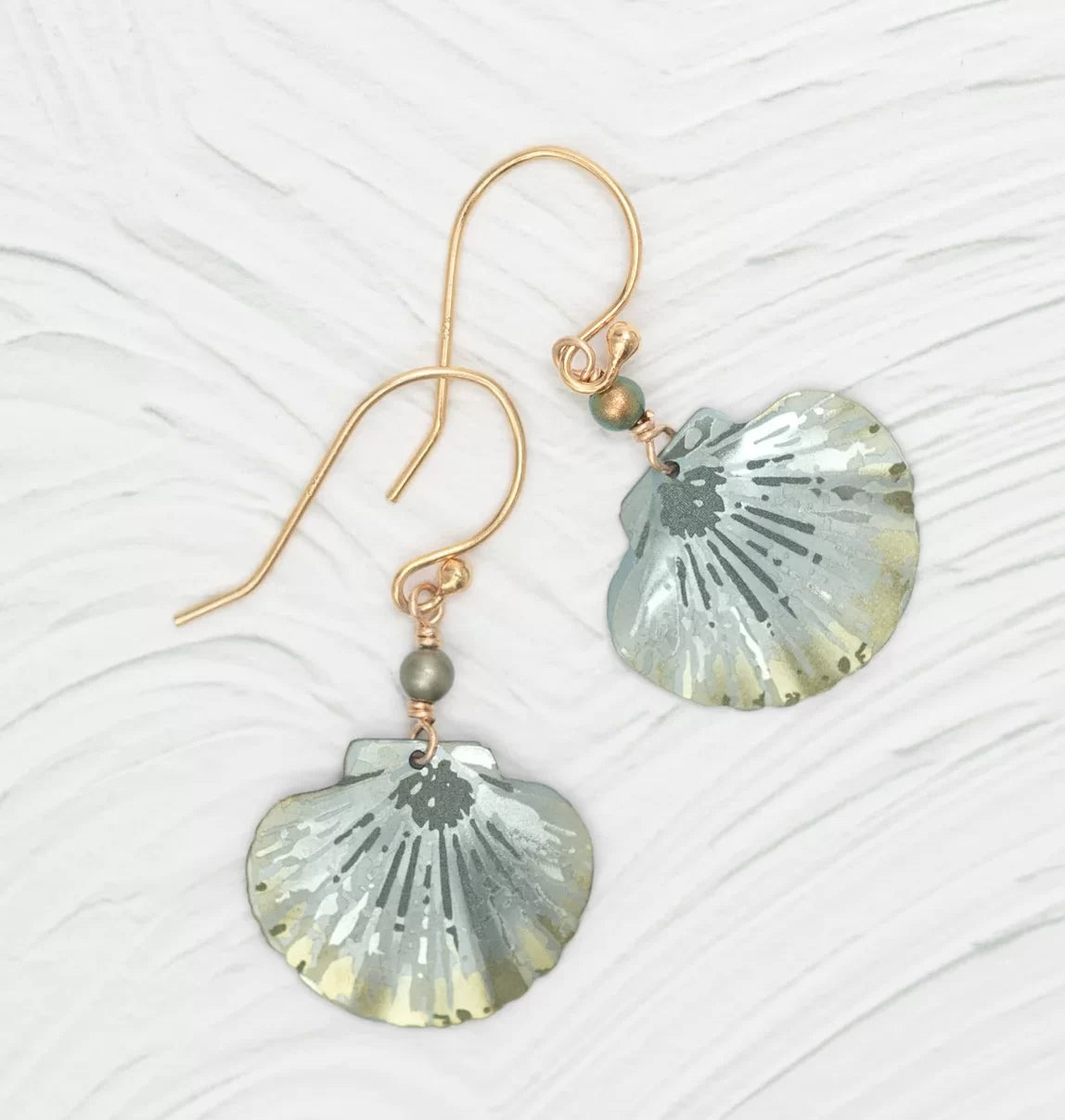 The Shelby Sage and Gold Earrings by Holly Yashi feature gold hook metallic scallop-shaped pendants with grey and gold hues, small bead accents, and Austrian crystal pearls. These hypo-allergenic earrings are beautifully displayed on a textured light background.