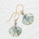 The Shelby Sage and Gold Earrings by Holly Yashi feature gold hook metallic scallop-shaped pendants with grey and gold hues, small bead accents, and Austrian crystal pearls. These hypo-allergenic earrings are beautifully displayed on a textured light background.