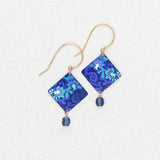 The Holly Yashi Garden Soiree Sapphire Blue Earrings are gold hook, hypoallergenic earrings with sapphire blue square floral pendants and a dangling blue bead, shown on a light textured background.