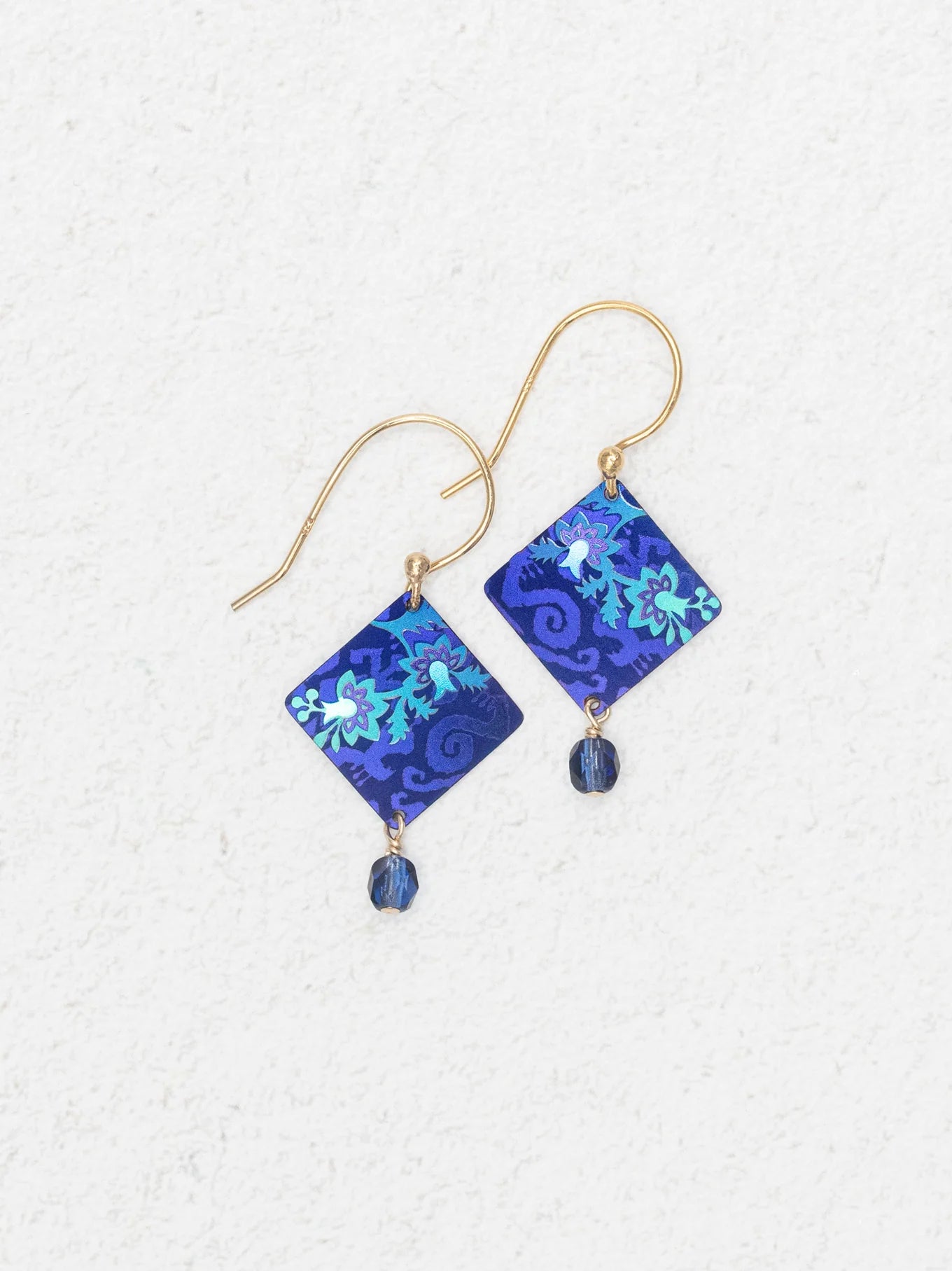 The Holly Yashi Garden Soiree Sapphire Blue Earrings are gold hook, hypoallergenic earrings with sapphire blue square floral pendants and a dangling blue bead, shown on a light textured background.