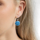 Close-up of a person with light hair wearing an earring. The design, resembling Gold Sandra Shore Sand Dollar Earrings from Holly Yashi Jewelry, features a delicate blue dandelion-like sphere on a thin hypo-allergenic metal hook. The softly blurred background enhances its elegance.