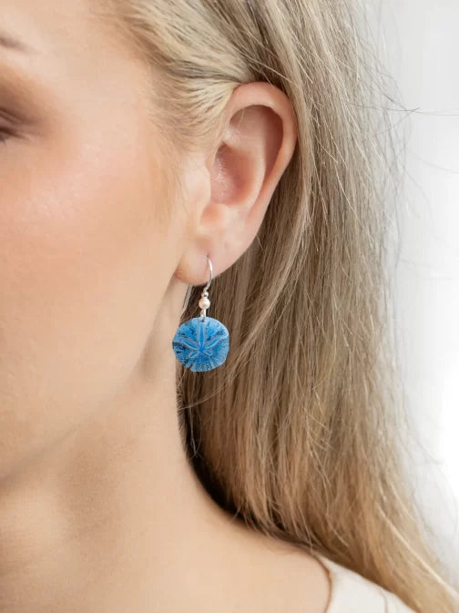 Close-up of a person with light hair wearing an earring. The design, resembling Gold Sandra Shore Sand Dollar Earrings from Holly Yashi Jewelry, features a delicate blue dandelion-like sphere on a thin hypo-allergenic metal hook. The softly blurred background enhances its elegance.