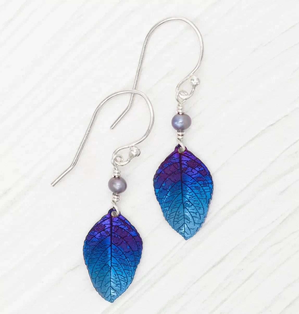 The Healing Leaf Navy Earrings by Holly Yashi feature iridescent purple and blue leaf-shaped pendants crafted from hypo-allergenic niobium, adorned with small gray beads and hanging from silver hooks, elegantly displayed on a light background.
