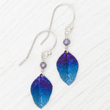 The Healing Leaf Navy Earrings by Holly Yashi feature iridescent purple and blue leaf-shaped pendants crafted from hypo-allergenic niobium, adorned with small gray beads and hanging from silver hooks, elegantly displayed on a light background.