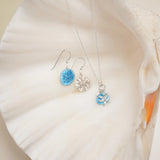 A silver necklace with a blue and white pendant and elegant Gold Sandra Shore Sand Dollar Earrings by Holly Yashi, featuring blue circular pendants, are beautifully displayed on a large, white seashell.