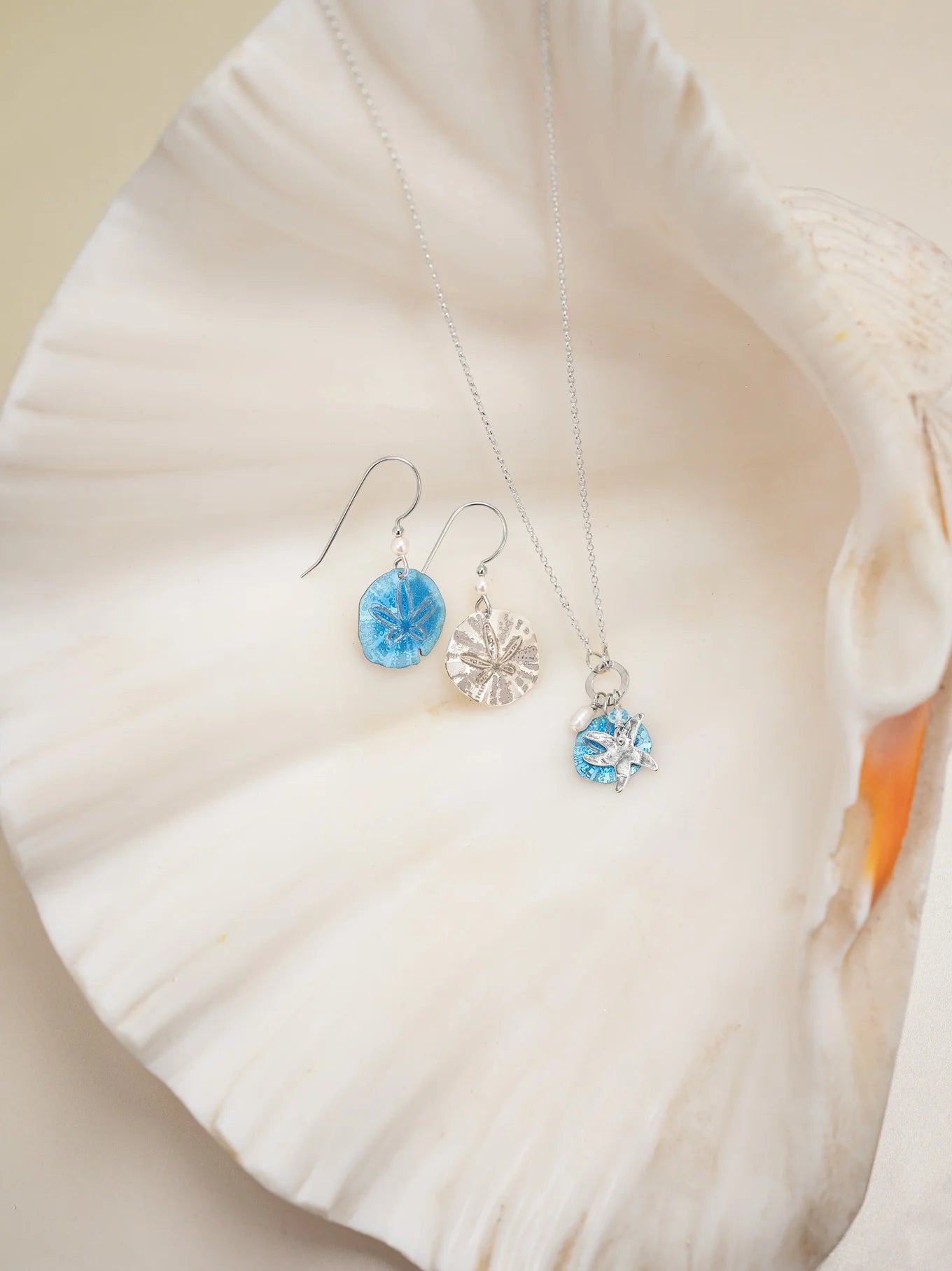 A silver necklace with a blue and white pendant and elegant Gold Sandra Shore Sand Dollar Earrings by Holly Yashi, featuring blue circular pendants, are beautifully displayed on a large, white seashell.