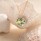 The Ocean Depths Teal Necklace from Holly Yashi Jewelry features a round gold pendant with turquoise and blue stones, elegantly draping over a white seashell. A shell-enhanced background adds to its coastal charm, reminiscent of bohemian glass artistry.