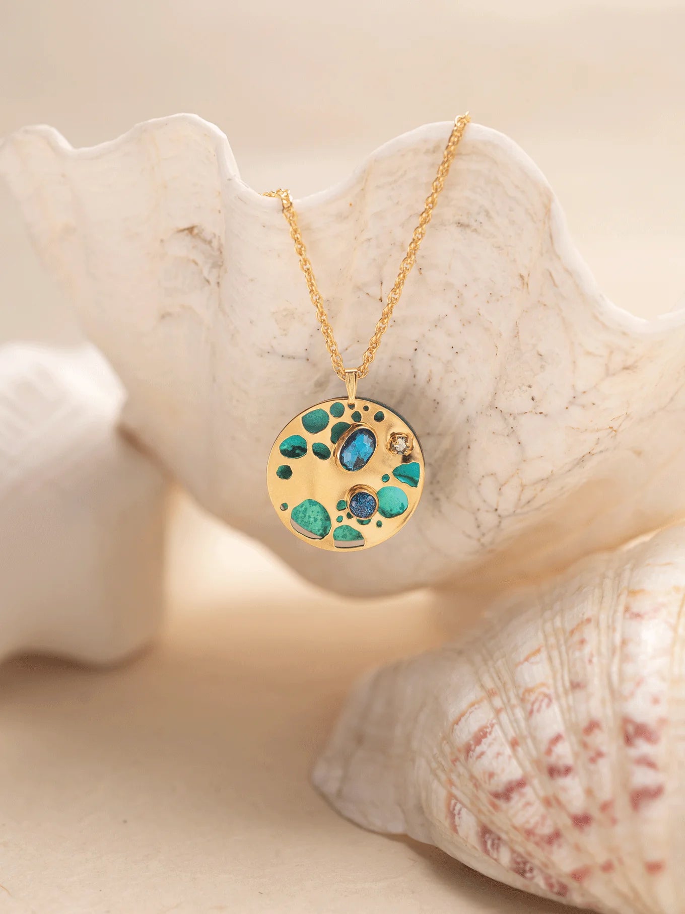 The Ocean Depths Teal Necklace from Holly Yashi Jewelry features a round gold pendant with turquoise and blue stones, elegantly draping over a white seashell. A shell-enhanced background adds to its coastal charm, reminiscent of bohemian glass artistry.