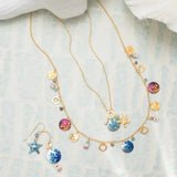 Hypo-allergenic gold necklaces and matching Holly Yashis Coral Reef Seashore Blue Earrings, enhanced with sea-themed starfish charms and colorful enamel discs, are elegantly displayed on a light blue textured surface with a white fabric draped above.