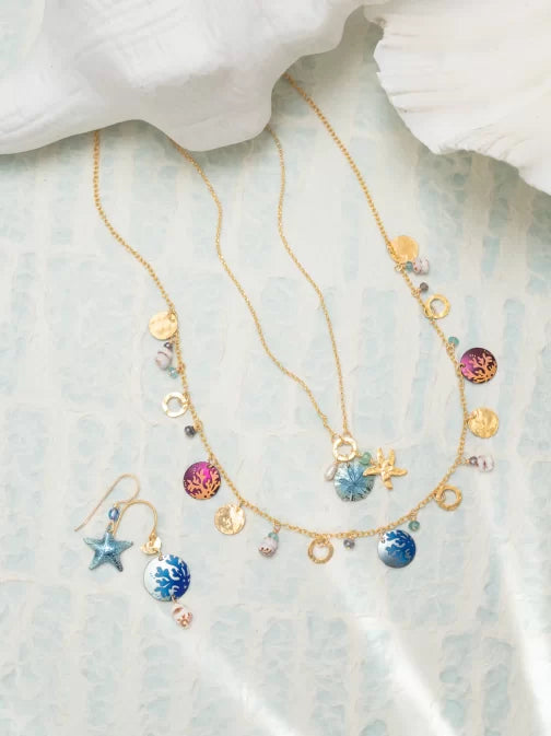 Hypo-allergenic gold necklaces and matching Holly Yashis Coral Reef Seashore Blue Earrings, enhanced with sea-themed starfish charms and colorful enamel discs, are elegantly displayed on a light blue textured surface with a white fabric draped above.